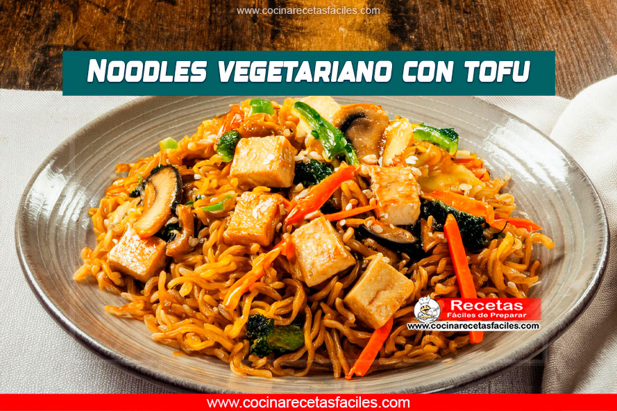 "Plato de noodles vegetarianos con tofu, zanahorias, pimiento rojo, calabacín y brotes de soja sobre una mesa de madera."