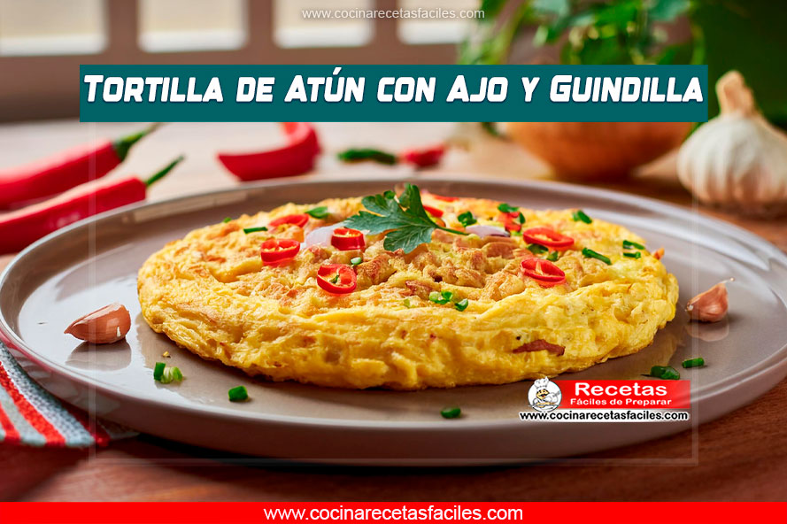 Tortilla de atún con guindilla roja y ajo, servida en un plato blanco sobre una mesa de madera con mantel, en el interior de una cocina con ventana.